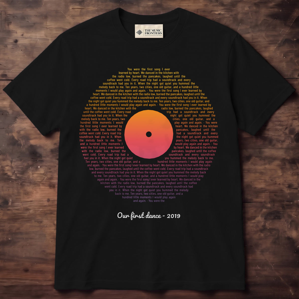 Black tee with lines of words filling a vinyl record in sunset gold, coral and purple, with a short line underneath